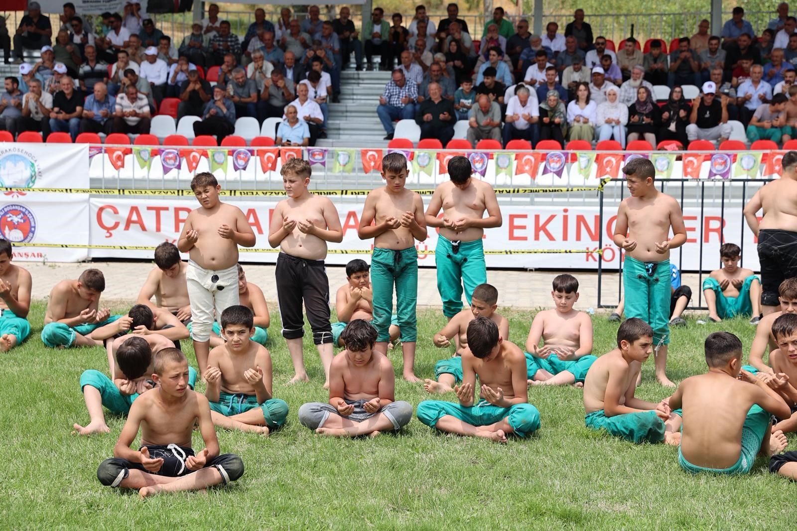 Çim alanda güreş kıyafetleri giymiş çocuklar otururken, tribünlerdeki kalabalık onları izliyor.