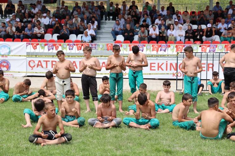 Çim alanda güreş kıyafetleri giymiş çocuklar otururken, tribünlerdeki kalabalık onları izliyor.