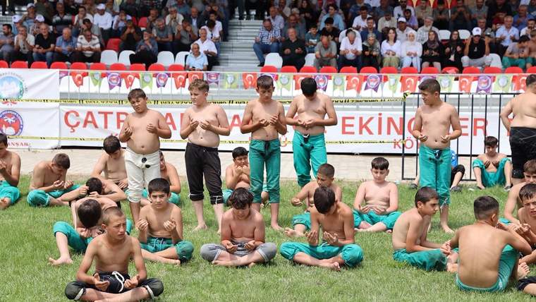 Çim alanda güreş kıyafetleri giymiş çocuklar otururken, tribünlerdeki kalabalık onları izliyor.