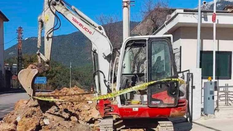Kırmızı beyaz bir ekskavatör, kazılmış toprak yığını ve sarı güvenlik şeridi yanında duruyor.