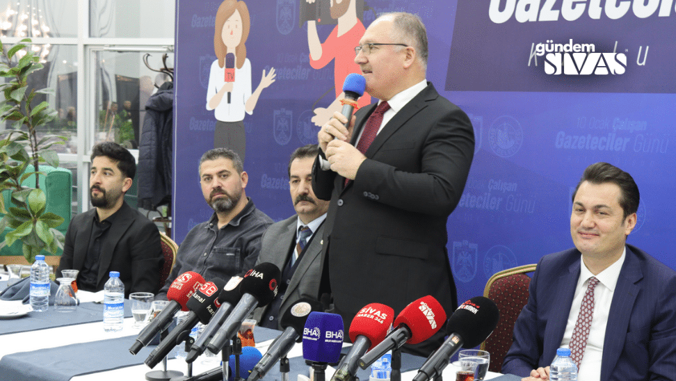 Bir kişi mikrofonla konuşurken, masada çeşitli medya logoları ve su şişeleri yer alıyor.