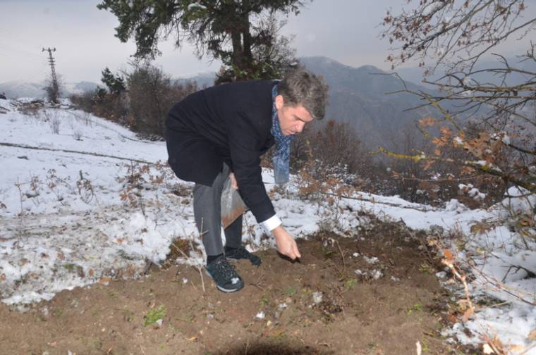 Karlı arazide çömelmiş bir kişi, elindeki tohumu toprağa bırakırken görülüyor.