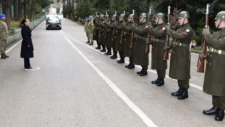 Askeri kıyafet giymiş askerler, selam durarak bir kadına karşı dizilmiş. Arka planda ağaçlar ve bir araç görünmekte.