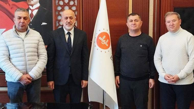Ahşap panelli bir odada dört erkek, üzerinde amblem bulunan beyaz bir flamanın önünde yan yana duruyor.