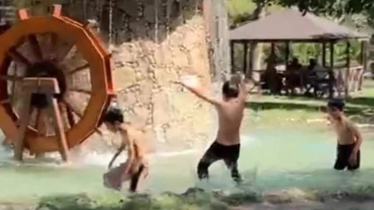 Su kenarında oynayan üç çocuk, arka planda su değirmeni görünmekte.