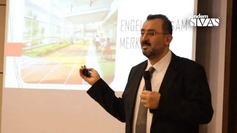 Takım elbiseli bir kişi, elinde mikrofon ve kumandayla ekrandaki Engelsiz Yaşam Merkezi sunumunu anlatıyor.