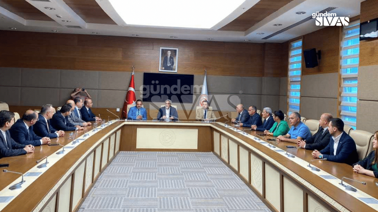 Toplantı odasında, masanın etrafında oturan kalabalık bir grup insan, önlerinde bir konuşmacı var.