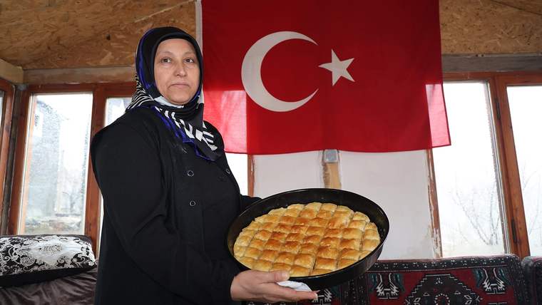 Başörtülü bir kadın, arkasındaki Türk bayrağı önünde dilimlenmiş baklava tepsisi tutuyor.