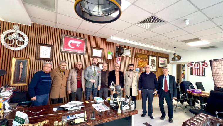 Ahşap panelli bir ofiste, masanın arkasında duran dokuz kişi ve öndeki kupalar görülüyor.