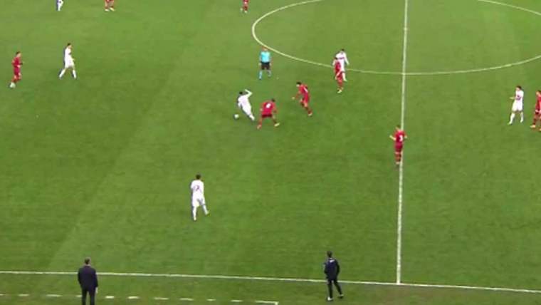 Futbol sahasında, beyaz formalı bir oyuncu topu kontrol etmeye çalışırken, kırmızı formalı iki oyuncu ona müdahale ediyor.