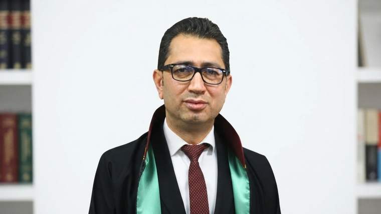 Gözlüklü bir adam, siyah cübbe ve kravatla kitaplıkların önünde kameraya bakıyor.
