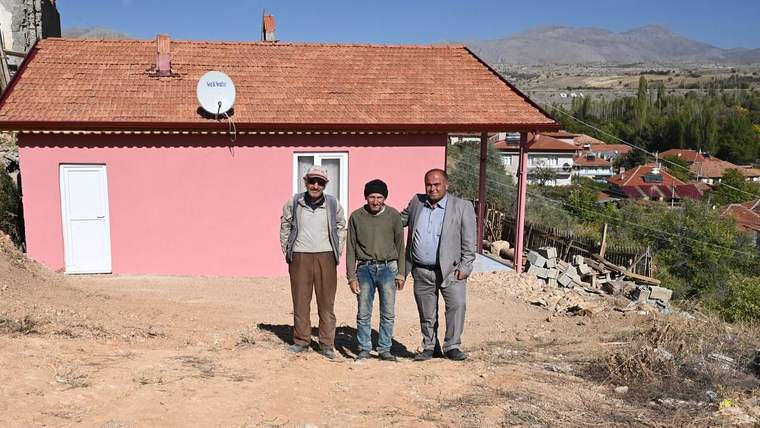 Pembe duvarlı, kiremit çatılı bir evin önünde duran üç erkek ve arkada dağlık yerleşim görülüyor.