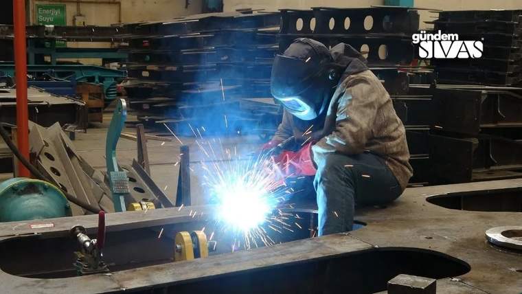 Atölyede koruyucu maskeli bir işçi, metal parçayı kaynak yaparken çevreye kıvılcımlar saçılıyor.