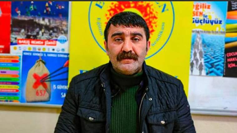 Bıyıklı bir erkek, siyah mont ve yeşil kazağıyla sarı arka plan ve afişlerin önünde duruyor.
