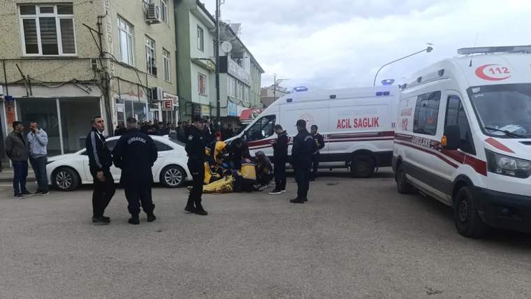 Bir grup polis ve sağlık çalışanı, yerde yatan bir kişiye müdahale ediyor; arka planda ambulanslar var.