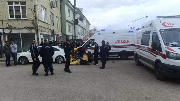 Bir grup polis ve sağlık çalışanı, yerde yatan bir kişiye müdahale ediyor; arka planda ambulanslar var.