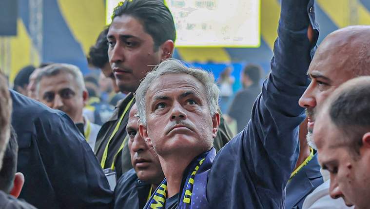 Gri saçlı bir adam, boynunda sarı-lacivert atkıyla kalabalık arasından kolunu kaldırıyor.
