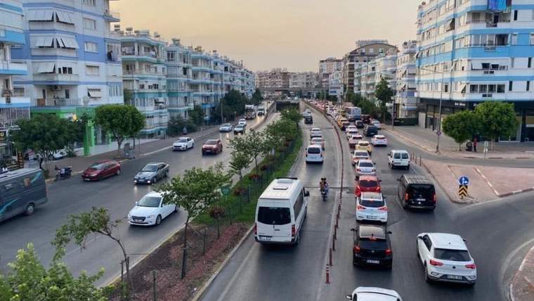 Bir cadde üzerinde araçların yoğun şekilde ilerlediği, apartmanların sıralandığı bir manzara.