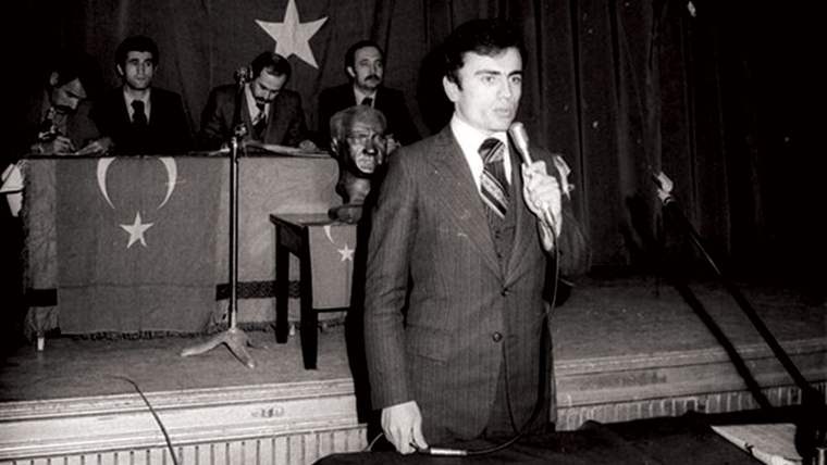 Bir adam, sahnede mikrofonla konuşuyor; arka planda Türk bayrağı ve bir heykel var.