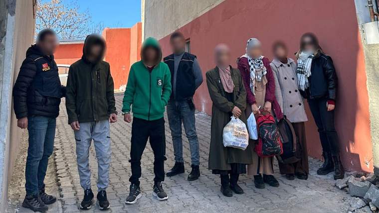 Bir duvar önünde yüzleri bulanıklaştırılmış yedi kişi, çantalarıyla birlikte yan yana duruyor.