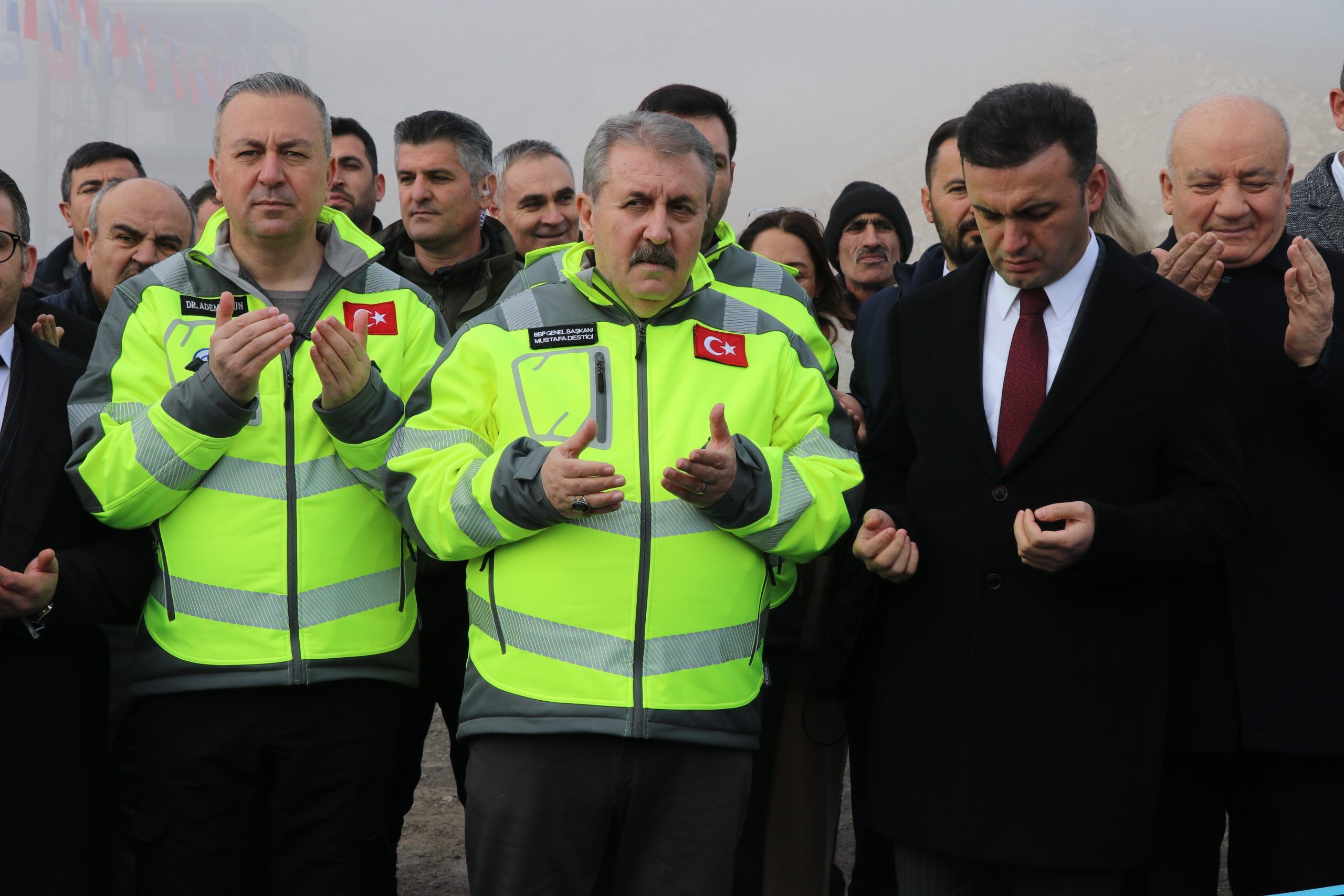 Açılış töreninde, yeşil yelek giymiş üç kişi dua ediyor, arka planda kalabalık bir grup yer alıyor.