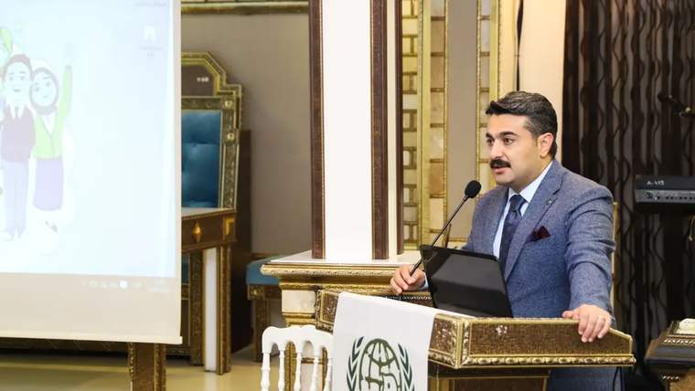 Kürsüde konuşan bir kişi, önündeki dizüstü bilgisayarla salondaki ekrana yansıtılan sunumun yanında duruyor.