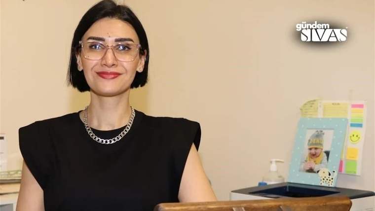 Gülümseyen bir kadın, masanın arkasında oturuyor; masada çocuk fotoğrafı ve çeşitli nesneler var.