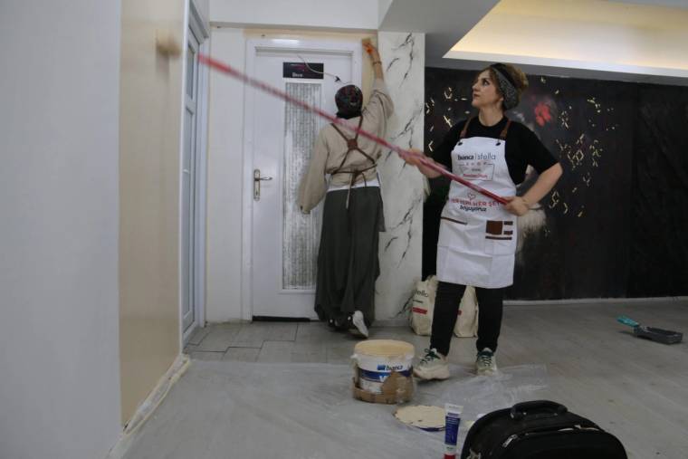 İç mekânda iki kadın çalışıyor; biri duvarı boyarken diğeri boya kovaları ve ekipmanların yanında duruyor.
