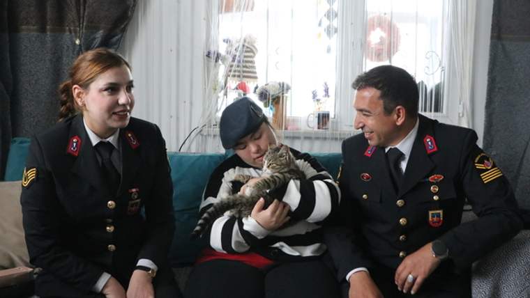 İki üniformalı kişi, kolunda kedi tutan genç kadının yanında ev ortamında oturuyor.