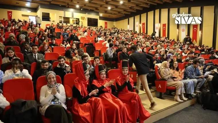 Konferans salonunda kalabalık izleyici topluluğu otururken, önde kırmızı geleneksel kıyafetli üç kişi görülüyor.