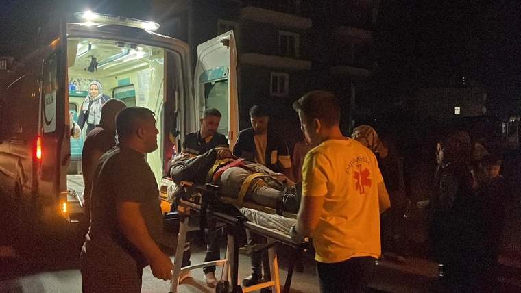 Ambulansın arkasında, sedye üzerinde bir yaralı taşınıyor; etrafında birkaç kişi var.
