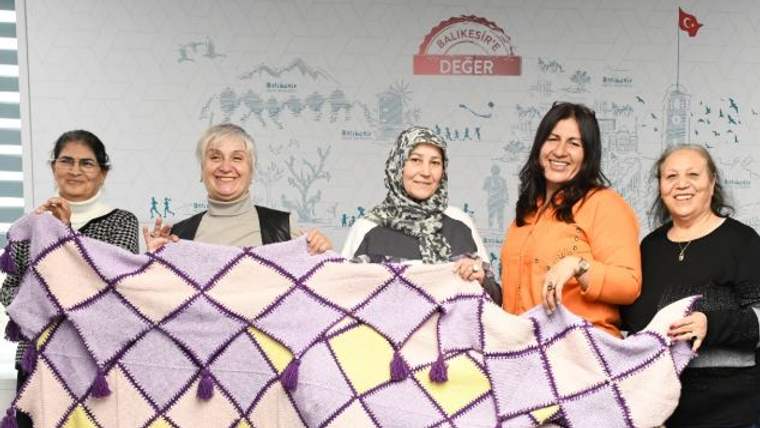 Beş kadın, mor ve sarı karelerden oluşan püsküllü el örgüsü battaniyeyi birlikte tutuyor.