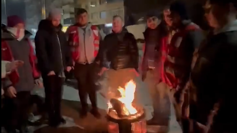 Kış gecesinde bir grup insan, ateşin etrafında toplanmış, sıcak bir ortamda sohbet ediyor.