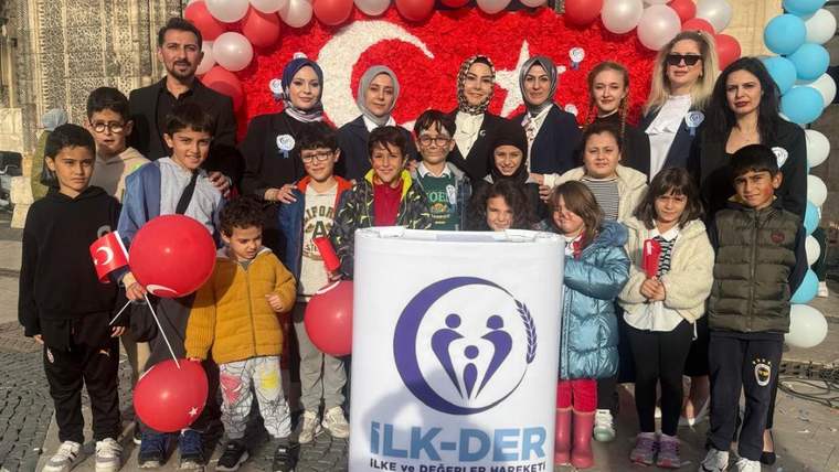 Çocuklar ve yetişkinler, İlk-Der'in düzenlediği etkinlikte bir araya gelmiş, balonlar ve Türk bayrakları tutuyorlar.