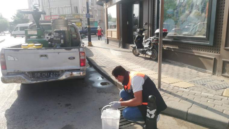Turuncu yelekli bir görevli, kaldırım kenarında beyaz kovayla çalışırken açık kasa araç yanında duruyor.