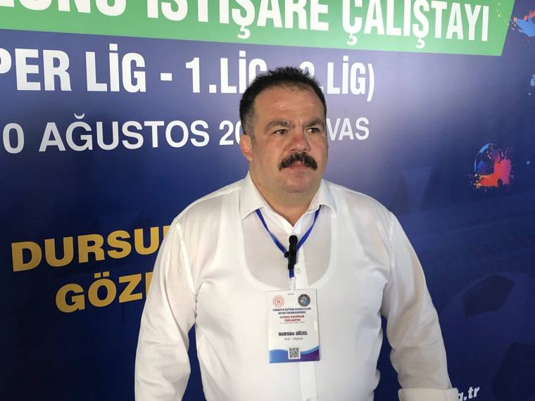 Bıyıklı bir kişi, boynunda yaka kartı ve mikrofonla yazılı bir etkinlik panosu önünde duruyor.