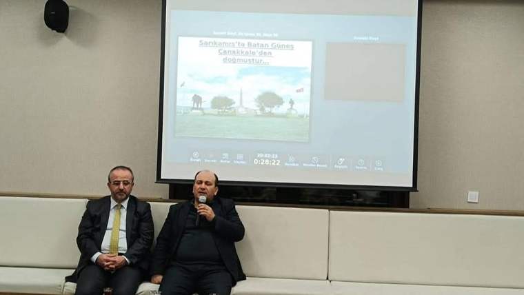 Konferans salonunda iki erkek oturuyor; sağdaki elindeki mikrofonla konuşurken arkadaki perdede sunum açılıyor.