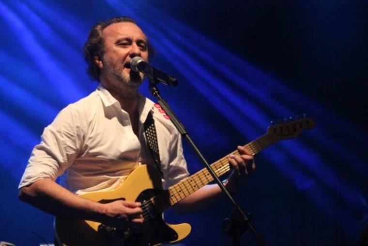 Beyaz gömlekli bir kişi, mavi ışıklarla aydınlanan sahnede gitar çalıp mikrofona şarkı söylüyor.