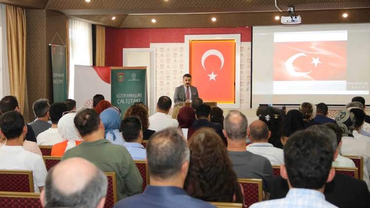 Toplantı salonunda kürsüde konuşan bir kişi, karşısındaki kalabalık ve Türk bayrağı eşliğinde görülüyor.