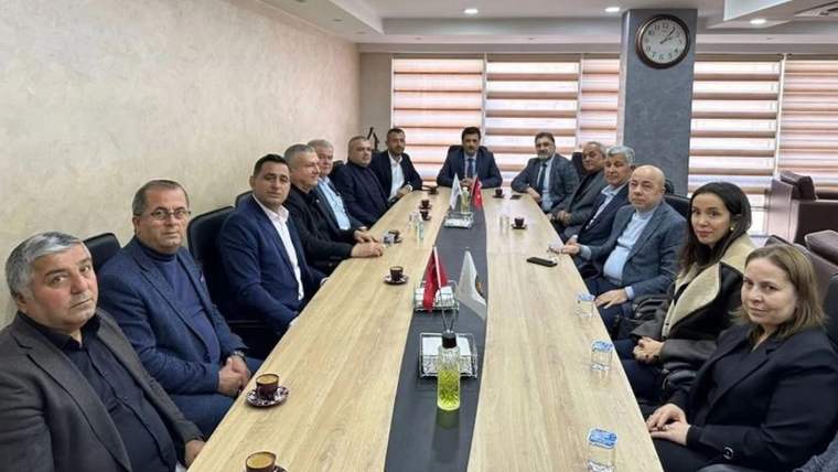 Toplantı masası çevresinde oturan bir grup kişi, masanın üzerinde bayraklar ve içeceklerle birlikte görülüyor.