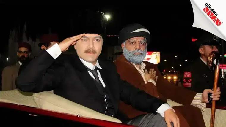 Siyah takım elbise giymiş bir kişi, selam durarak oturuyor; yanında başka bir kişi var.