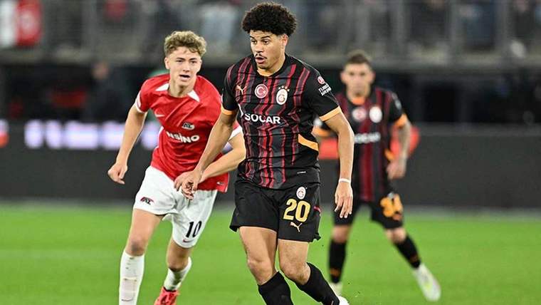 Siyah ve turuncu formasıyla bir futbolcu, top sürerken arkasında başka bir futbolcu yer alıyor.