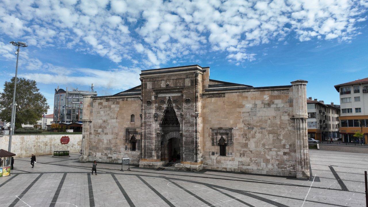 Taş cepheli tarihi bir yapı, geniş taş döşeli meydanda bulutlu mavi gökyüzü altında görülüyor.