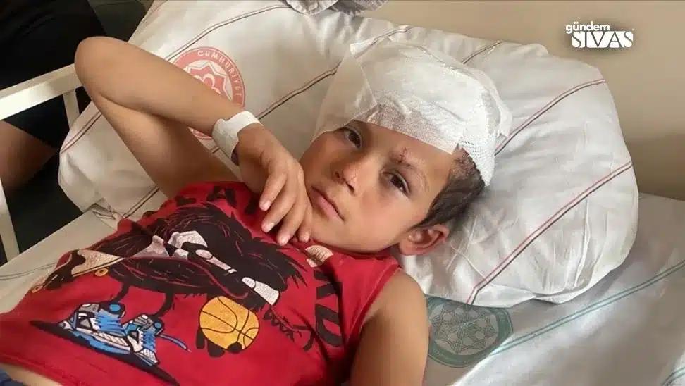 Başında beyaz bandaj bulunan bir çocuk, hastane yatağında yatarken elini yüzüne götürüyor.