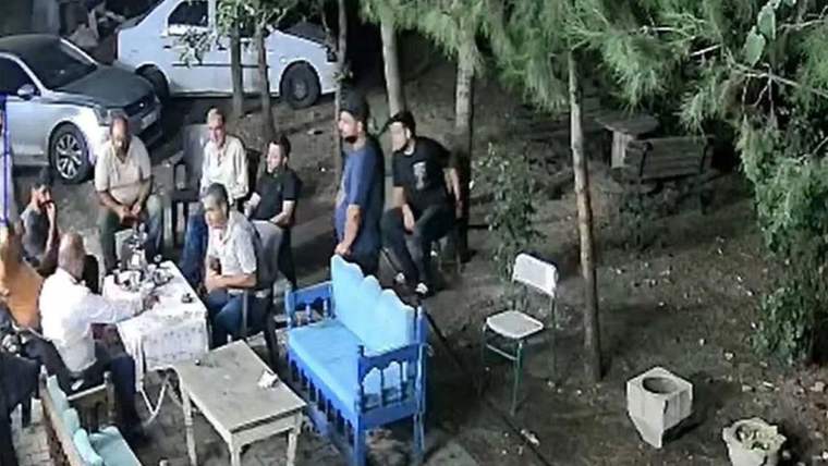Bir grup insan, açık alanda bir masanın etrafında oturuyor; arka planda ağaçlar ve bir araç görünmekte.