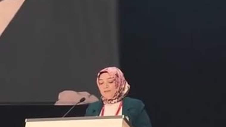 Başında eşarp bulunan bir kişi, mikrofonlu kürsüde önündeki metne bakarken görülüyor.