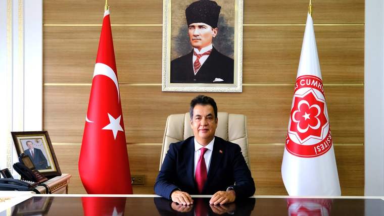 Makam odasında takım elbiseli bir kişi, Türk bayrağı ve üzerinde amblem bulunan beyaz bayrak arasında oturuyor.