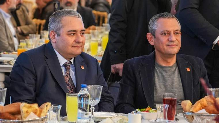 Kalabalık bir salonda masada oturan iki erkek, önlerindeki tabak ve içeceklerle birlikte görülüyor.