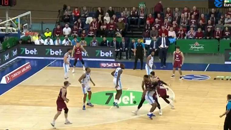 Basketbol maçında bordo ve beyaz formalar giyen oyuncular, salondaki seyirciler önünde mücadele ediyor.