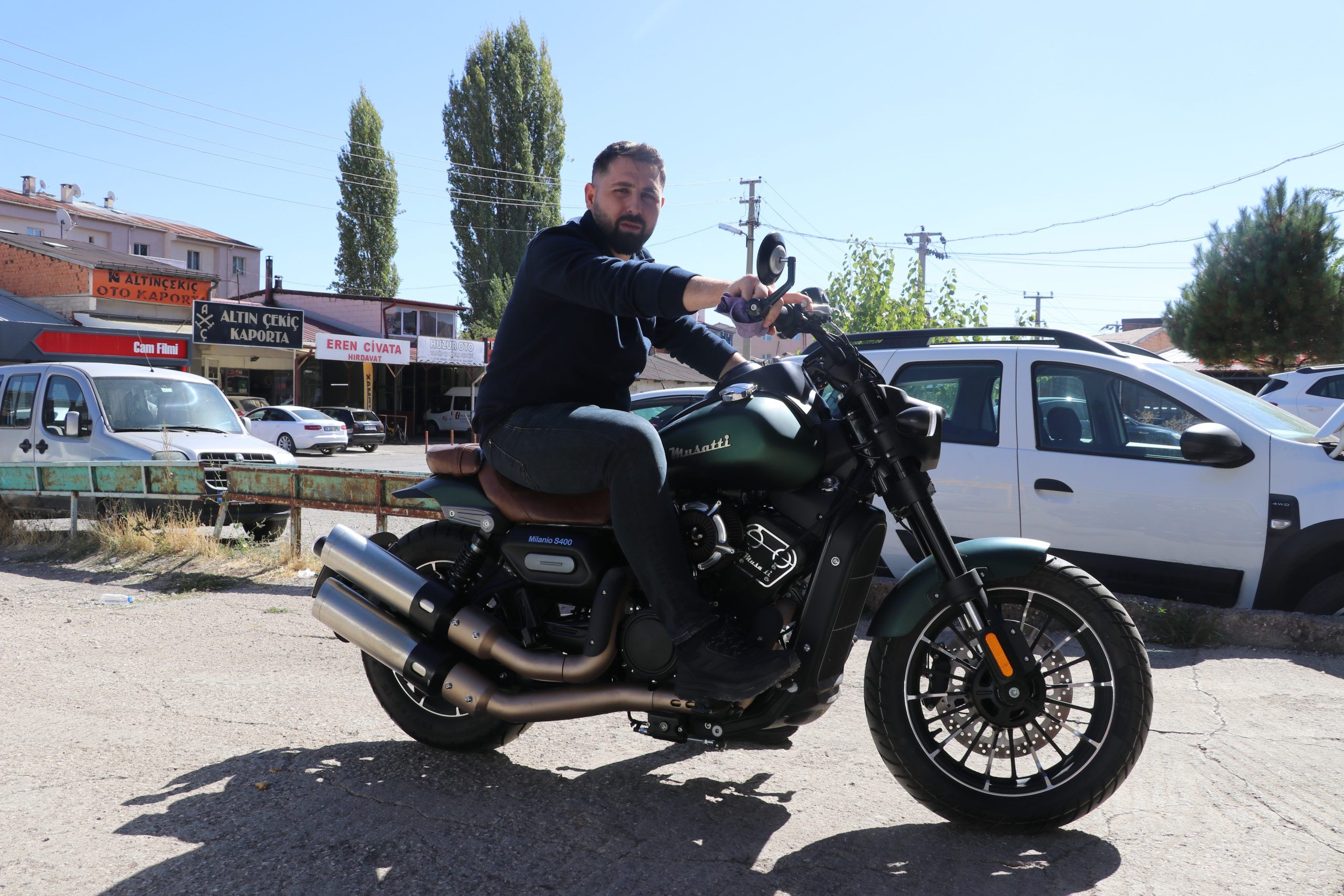 Motosiklet üzerinde oturan bir adam, arka planda park halindeki araçlar ve dükkanlar görünüyor.
